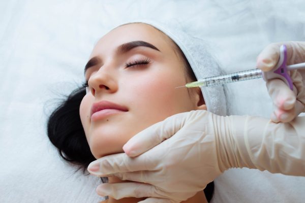 Smile lifting and lip augmentation. Beautician doctor hands doing beauty procedure to female face with syringe. Young woman's mouth contouring with filler injection. Marionette lines treatment.